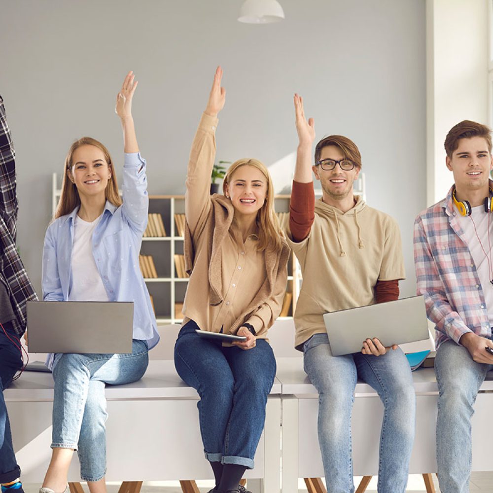 Group of happy smiling active students raising hands sitting on desk with digital devices. Young people agree with good idea or answer questions in lecture class. Studying at school or college concept