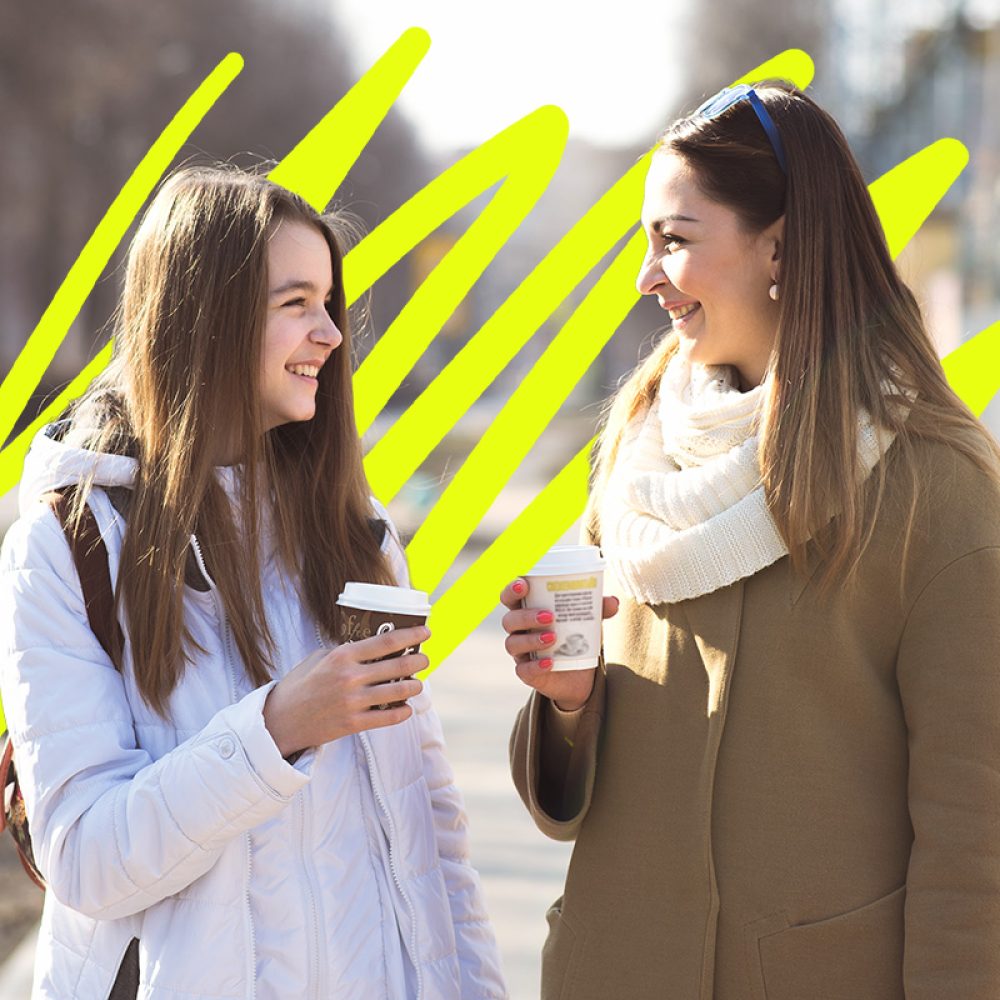 Mother and daughter talking, laughing and smiling on the street, drinking coffee in cups, in the fall or spring day, happy family.